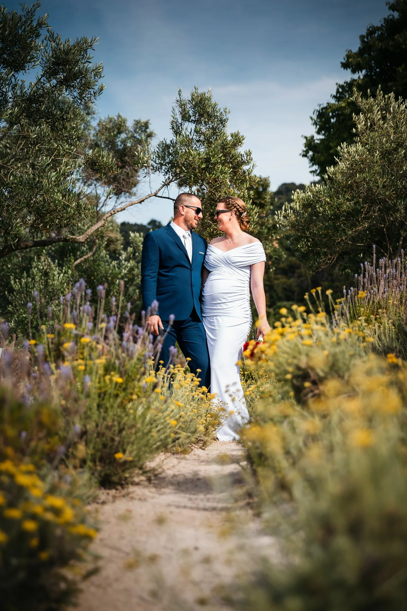A man and a woman in wedding attire walking through a garden with yellow and purple flowers, surrounded by trees and greenery.