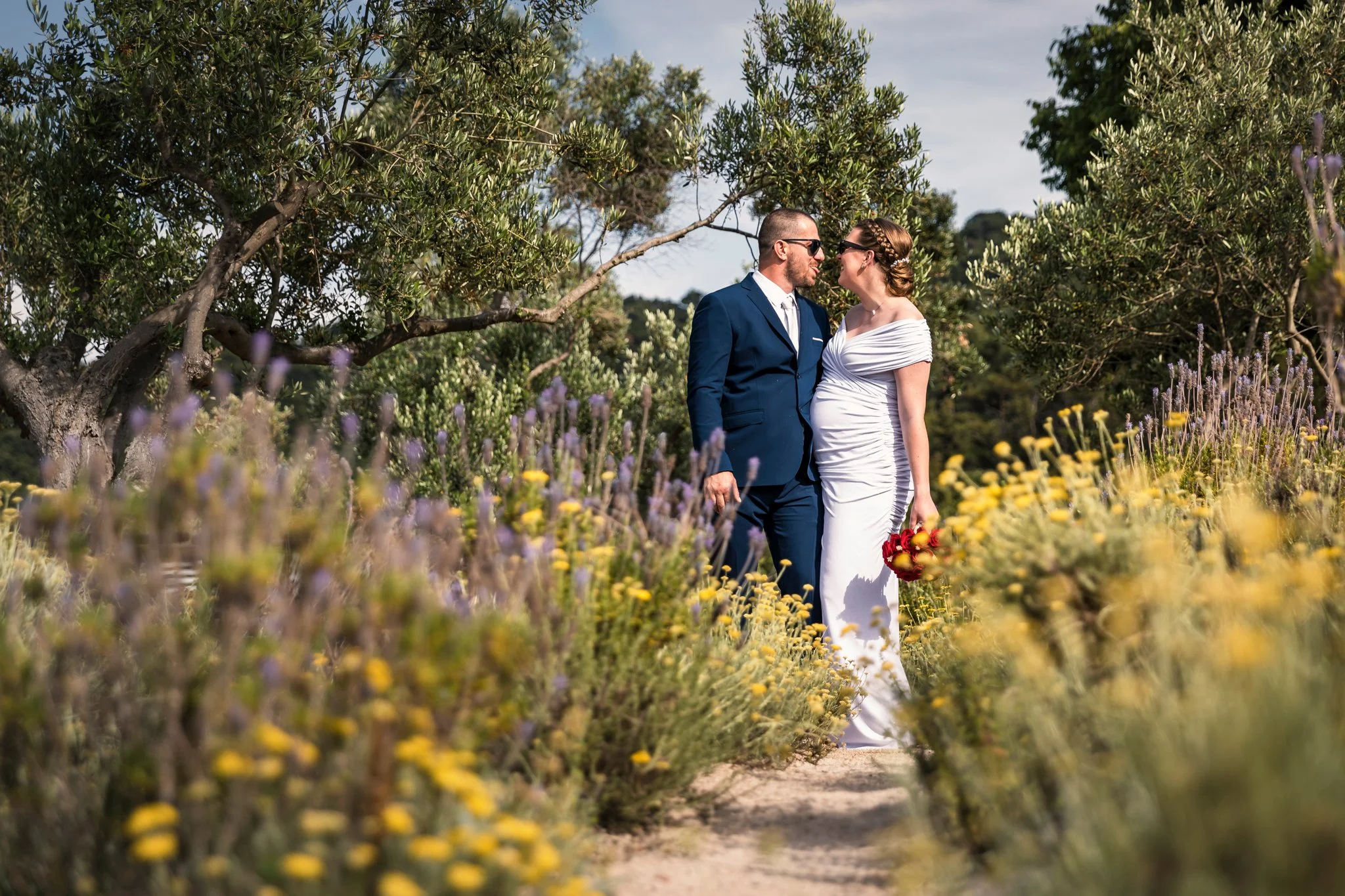 A bride and groom standing close together in a garden, gazing at each other, with trees and purple and yellow flowers surrounding them.