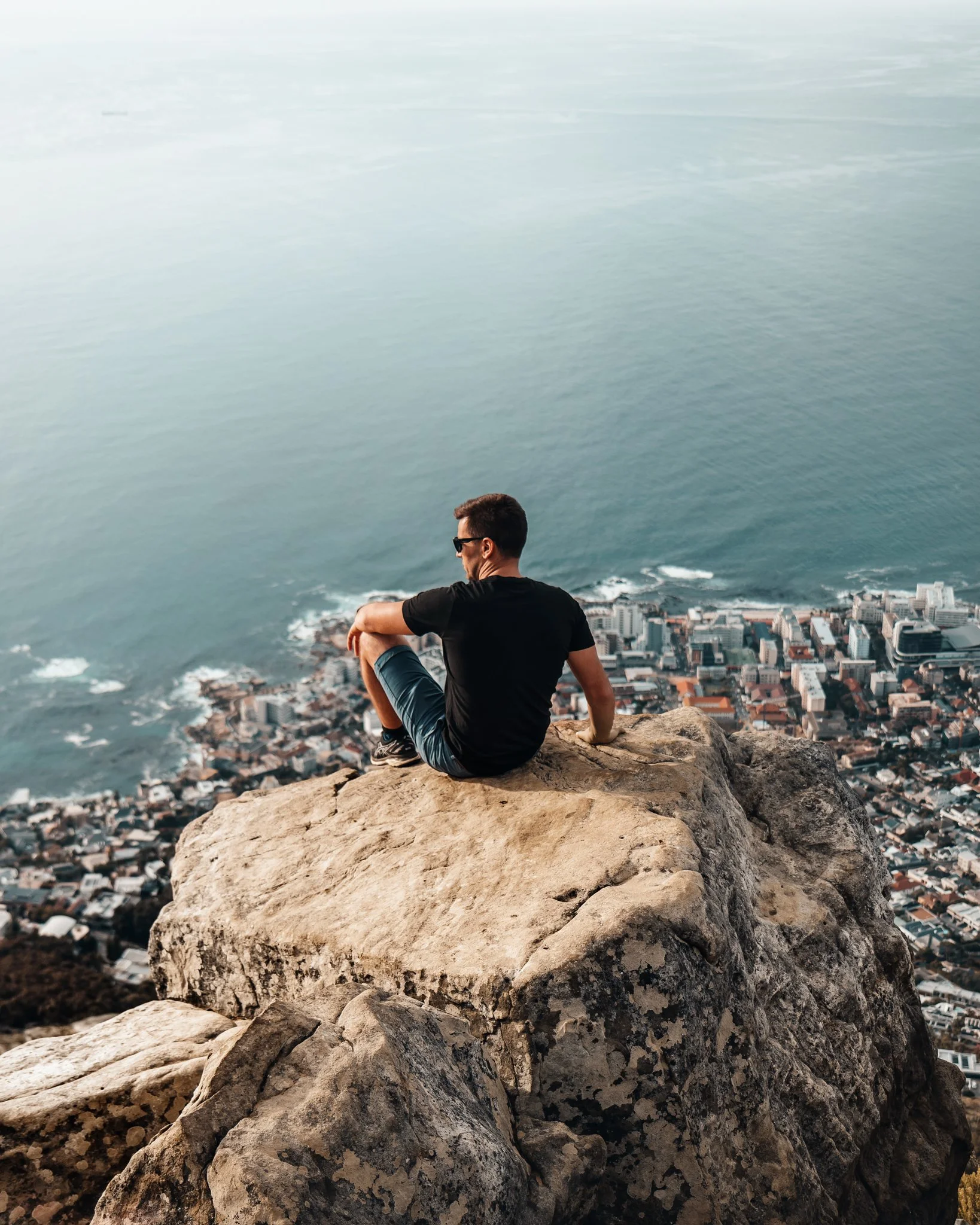 A man sitting on a large rock at the edge of a cliff overlooking a city and the ocean with ships in the distance.