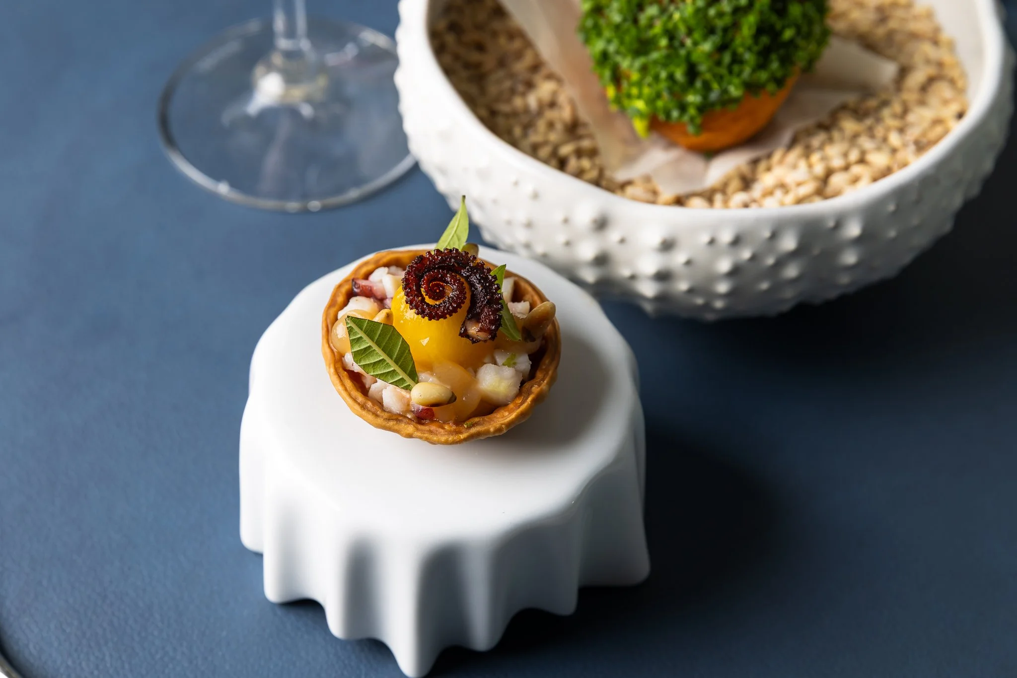A small tart filled with fruit and garnished with green leaves and a dark decorative element, placed on a white stand with a larger bowl of nuts and a wine glass in the background.