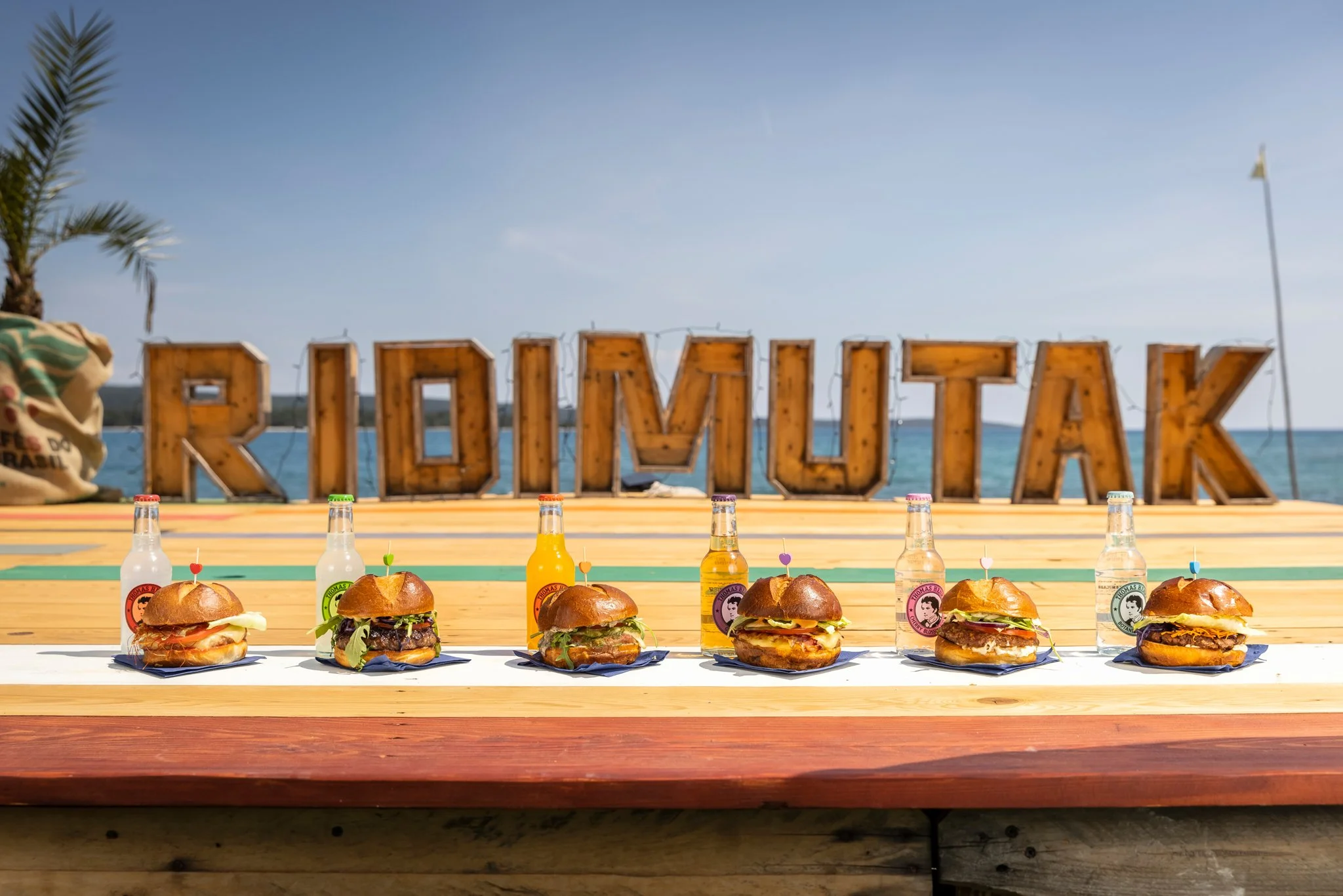 Display of six gourmet burgers with buns, lettuce, cheese, and toppings, lined up on a white surface in front of a wooden table, with colorful bottled drinks. In the background, a large wooden sign spelling 'REOVI' is set against a waterfront view, w