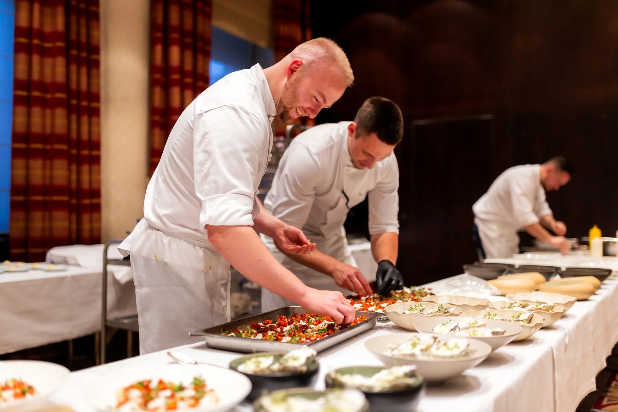 Chefs in white uniforms preparing and serving food at a banquet table with various dishes in a formal setting.