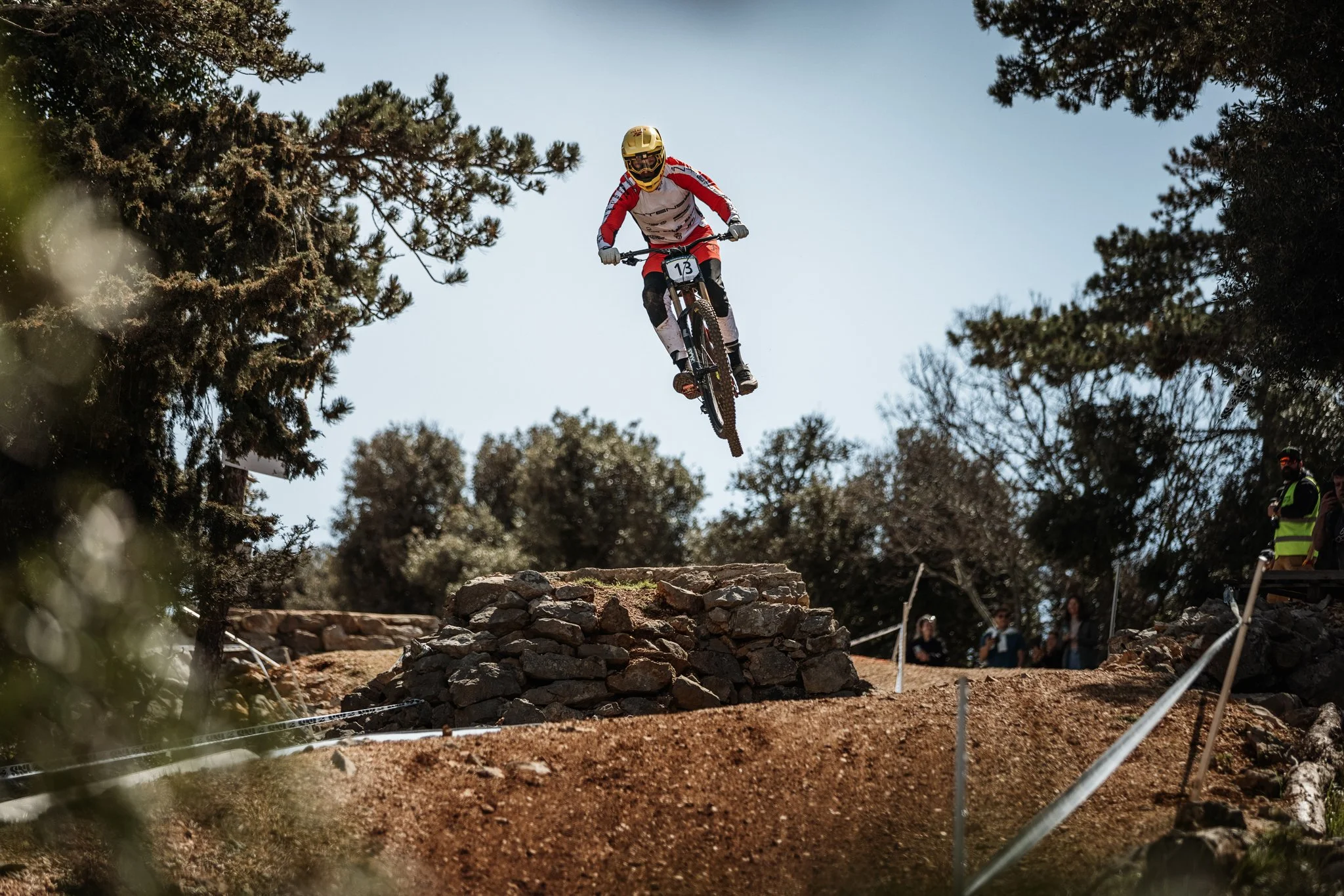 A mountain biker wearing a yellow helmet and red and white gear is riding mid-air over a rocky jump at an outdoor dirt trail, surrounded by trees and spectators.