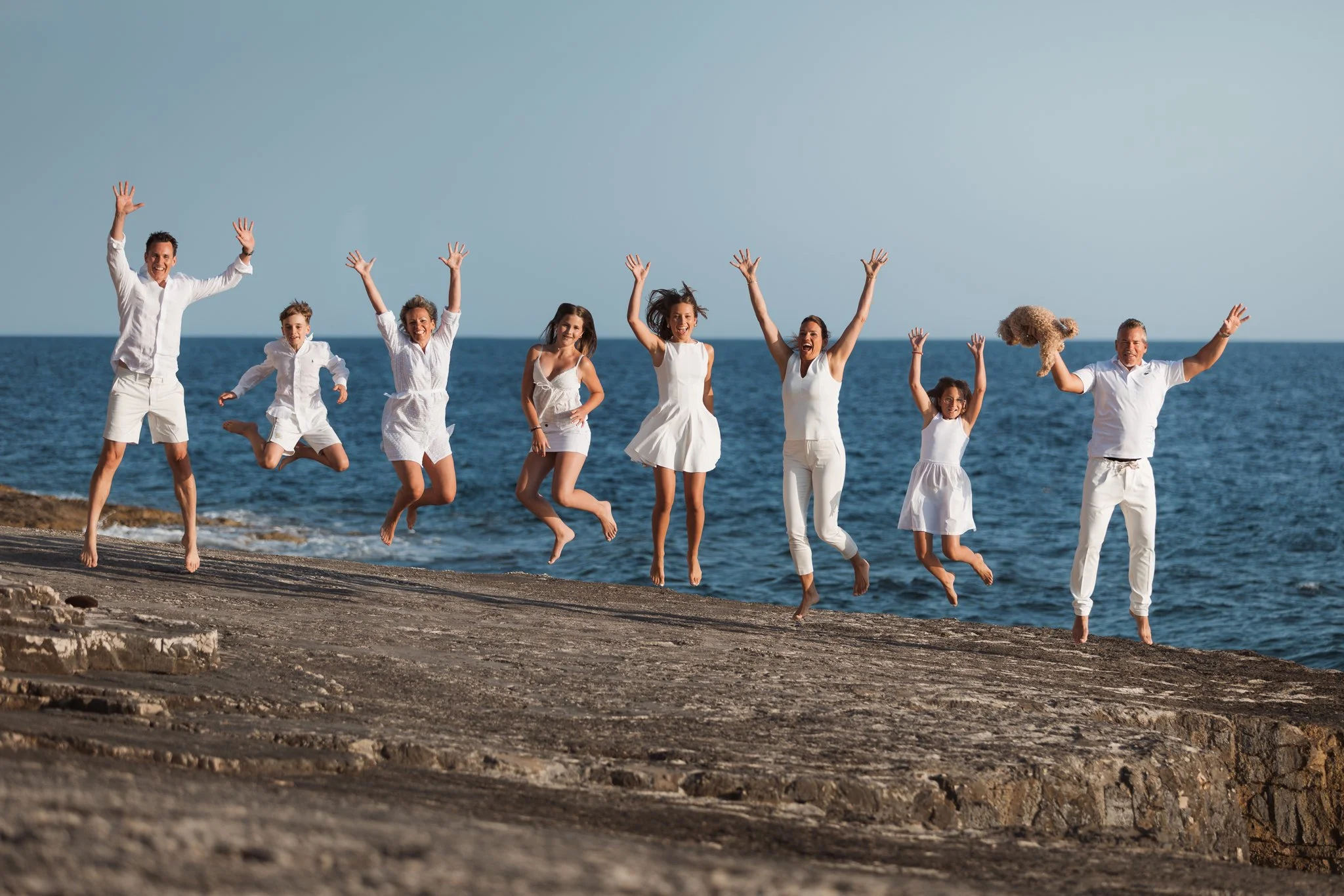 A group of seven people, including children and adults, jumping on a beach with the ocean in the background, all dressed in white and smiling.