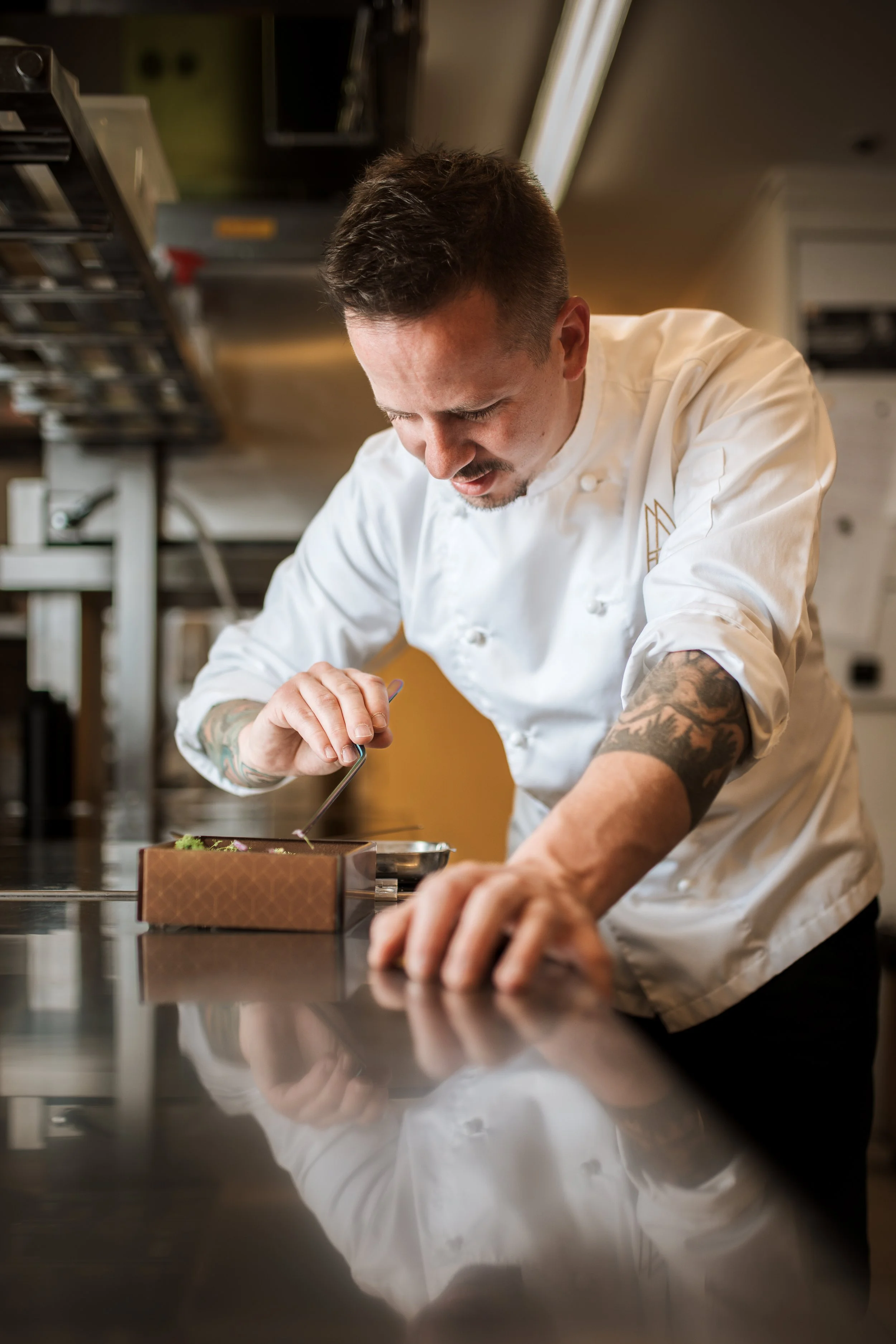 A professional chef with tattoos wearing a white chef's coat is carefully garnishing a dish in a commercial kitchen.