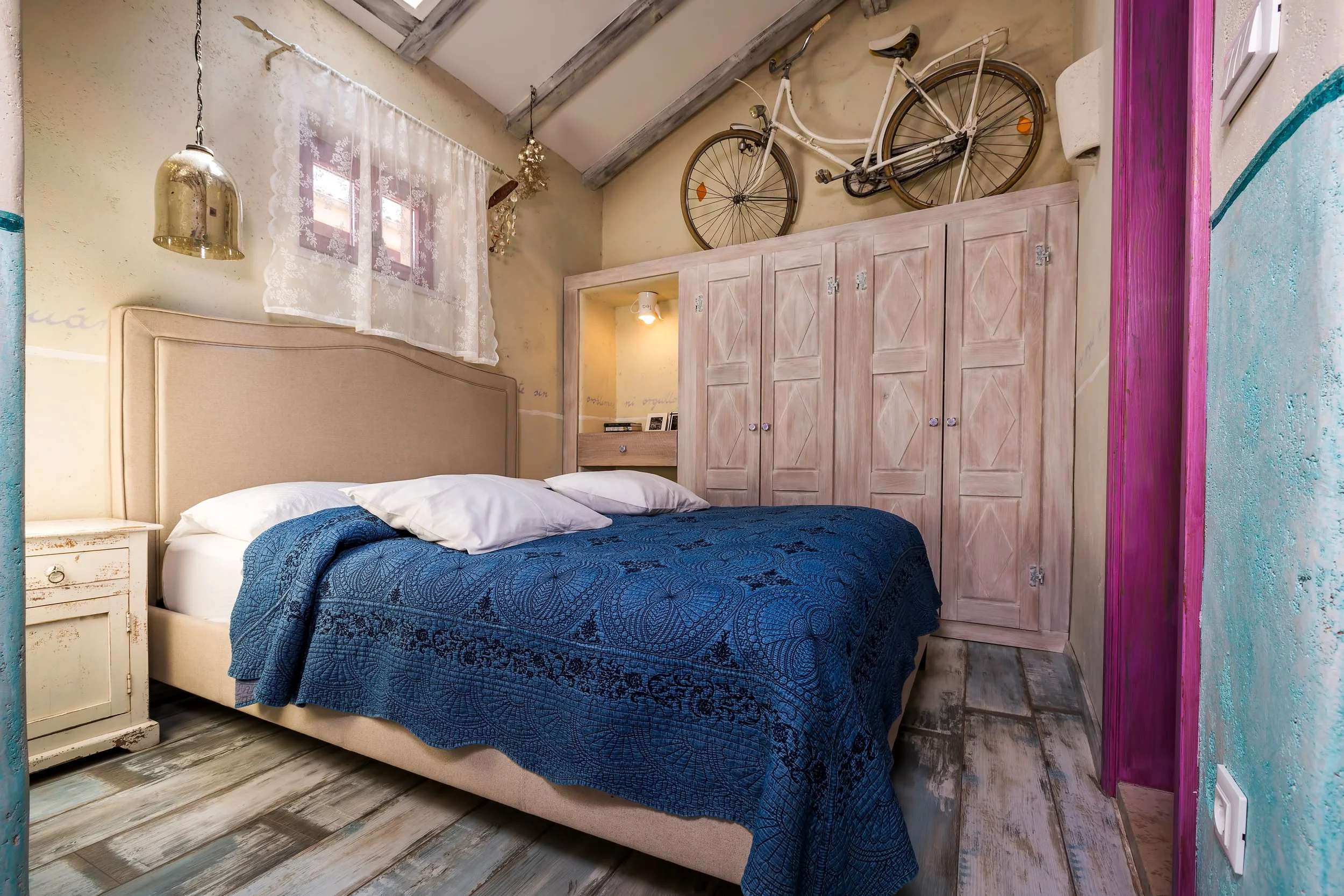 A cozy bedroom with a beige upholstered bed, blue patterned bedspread, and white pillows. There is a small off-white vintage nightstand with a drawer to the left of the bed. A white wardrobe with four doors is behind the bed, and a bicycle is mounted