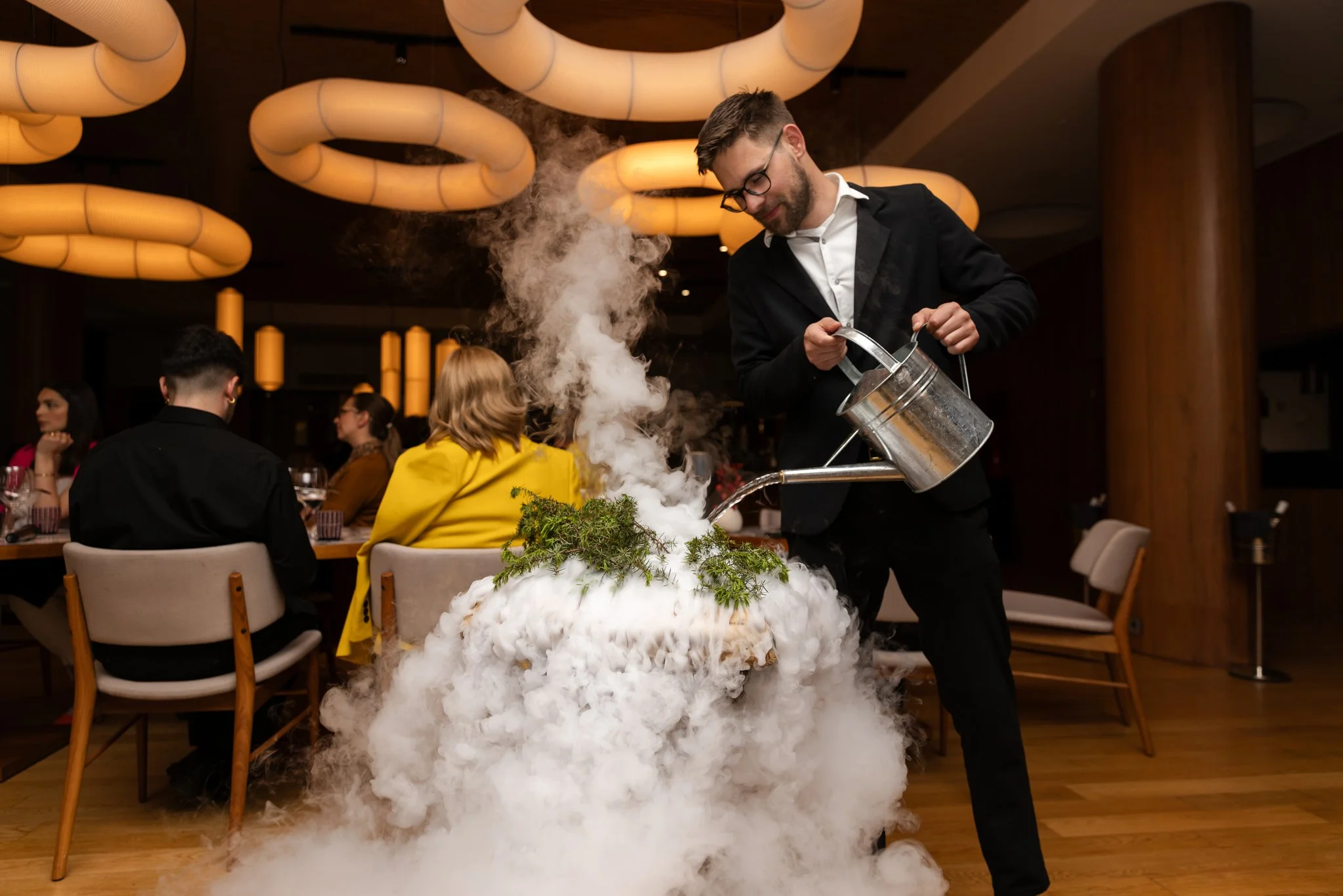 A man in a black suit creates a cloud of dry ice smoke at a restaurant table decorated with greenery, while a waiter pours liquid into it. The restaurant has modern lighting with large, circular, illuminated fixtures and several seated diners in the 