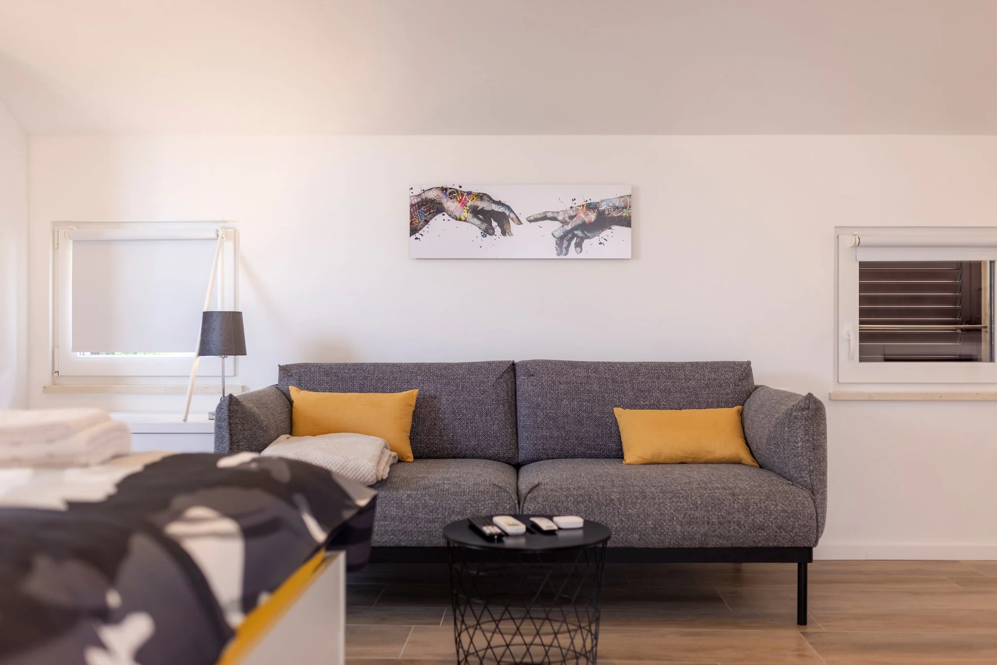 Modern living room with a gray sofa accented with yellow pillows, a black wire coffee table, and artwork of two hands reaching towards each other hung on a white wall between two windows.