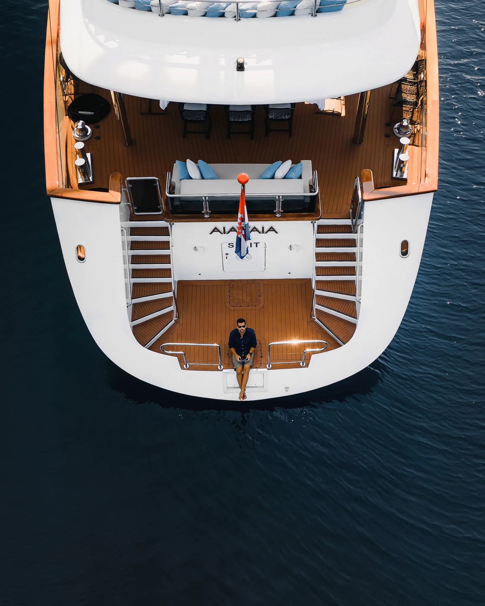 Top-down view of a yacht on the water, with a woman seated on the stern holding a remote control, and a man standing nearby. The yacht has a wooden deck, a white canopy, and a seating area with blue and white cushions.