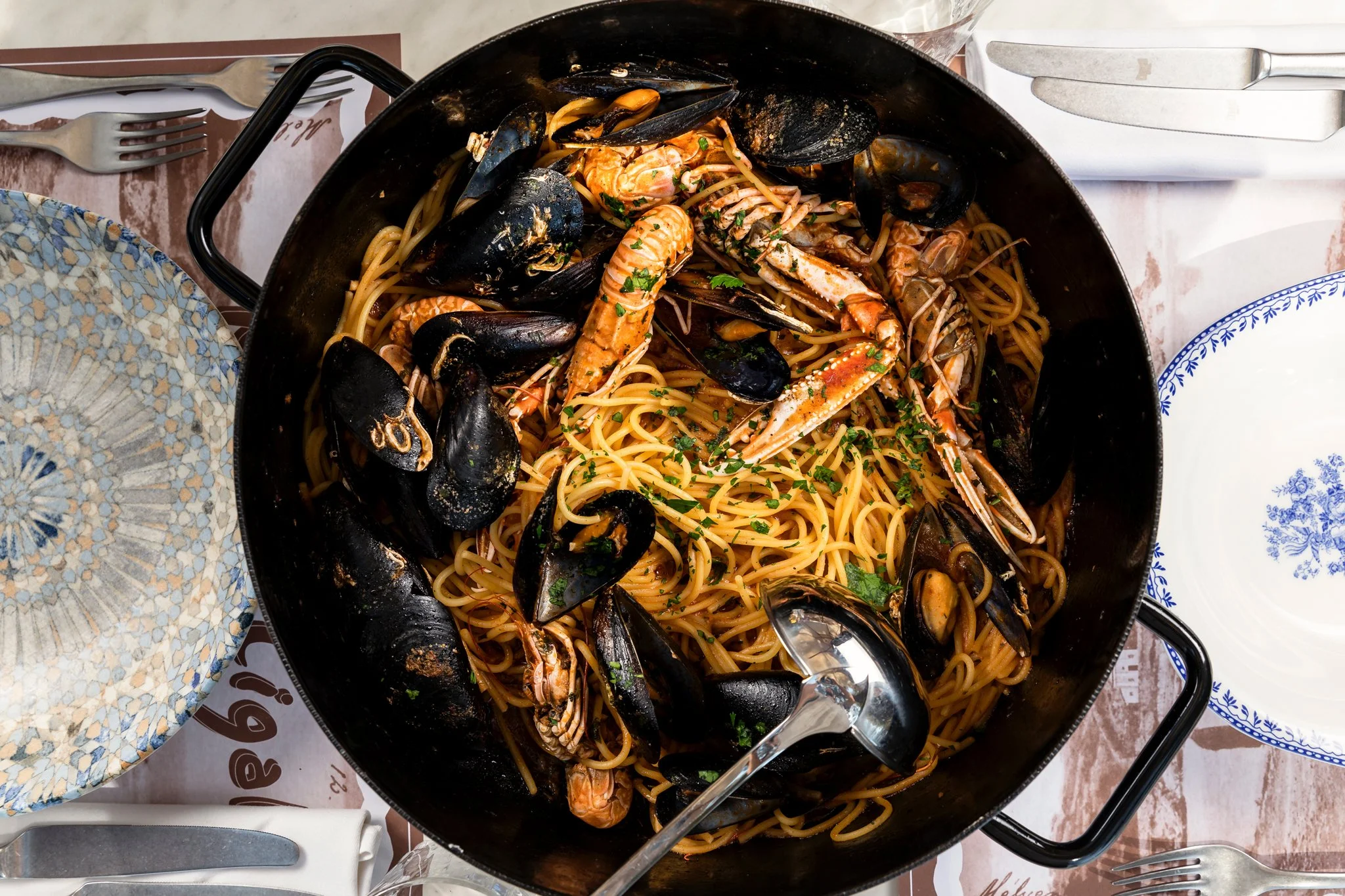 A large black skillet filled with seafood pasta, including mussels, shrimp, and crab, garnished with chopped parsley, on a table with plates, forks, and knives.