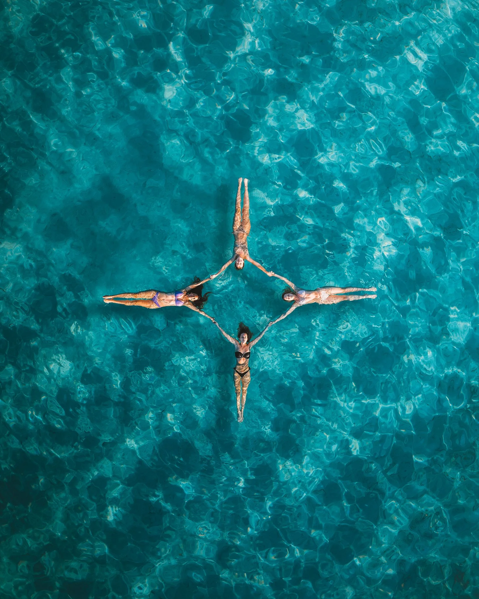 Four women in swimsuits holding hands in a circle while floating in a large, clear blue swimming pool.