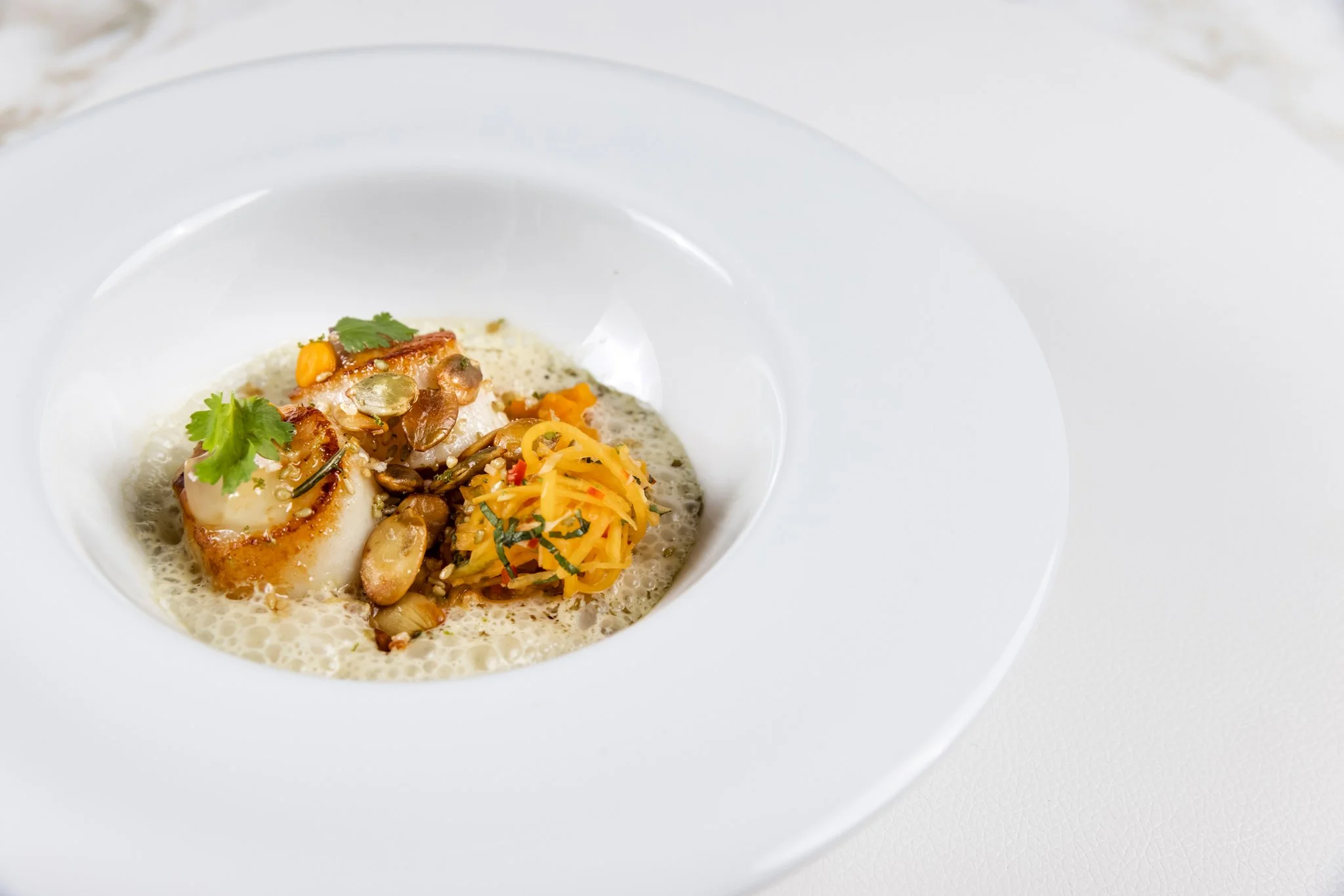 A white bowl with a small amount of soup, topped with vegetables, herbs, and possibly seafood.