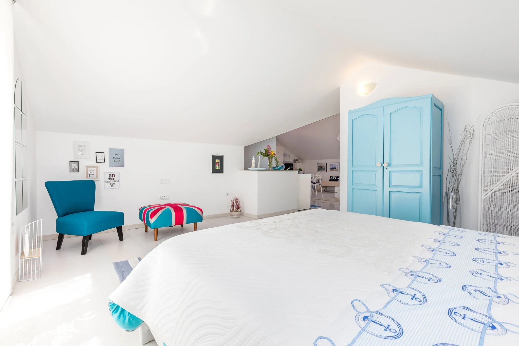 A bright bedroom with white walls and ceiling, featuring a large bed with white and blue embroidered bedding. There is a blue armchair and a colorful ottoman in front of it. On the wall behind, there are several framed artworks. A blue wardrobe is si