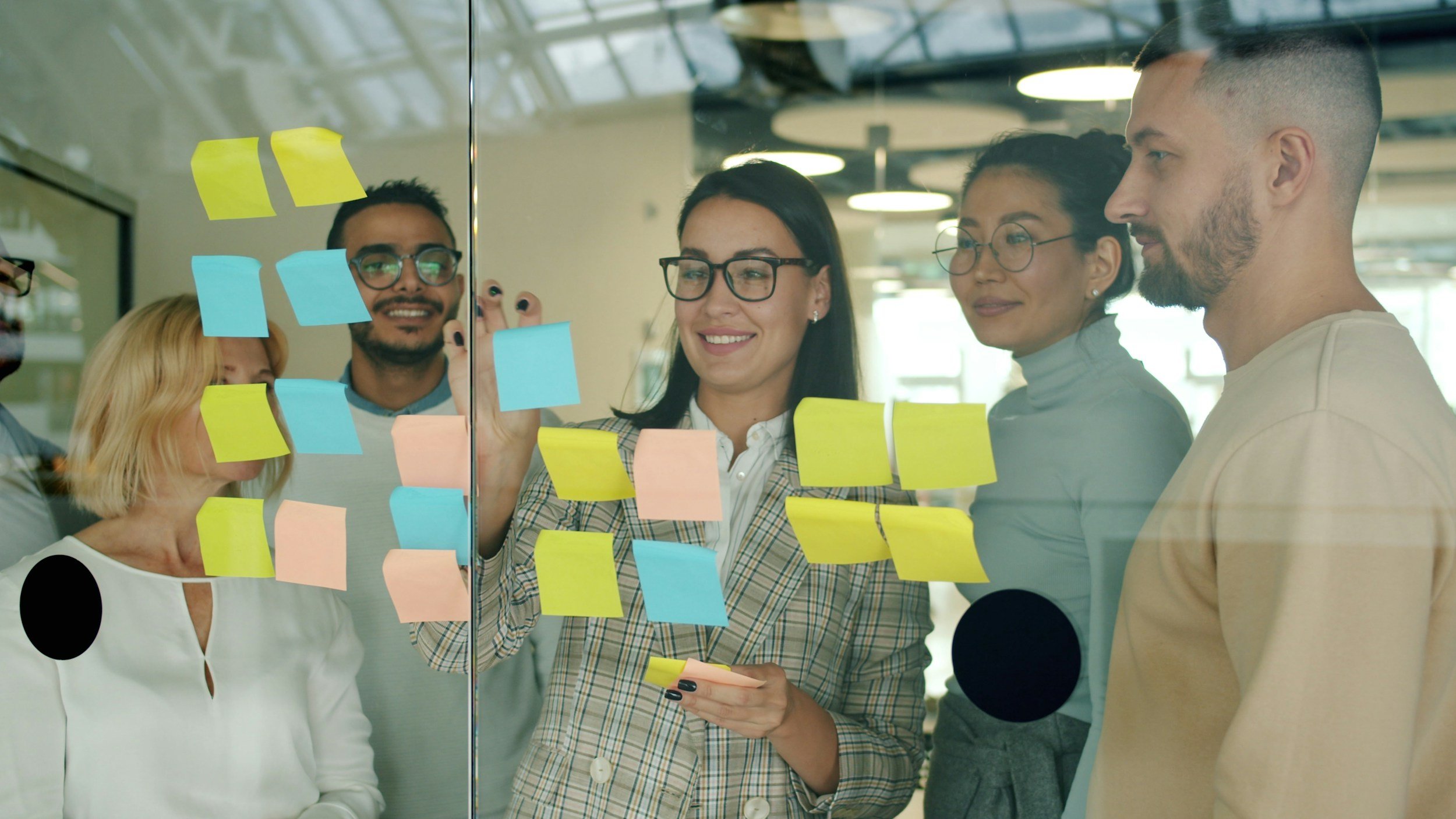 A group of five diverse people working together behind a glass wall covered with colorful sticky notes, in an office setting.