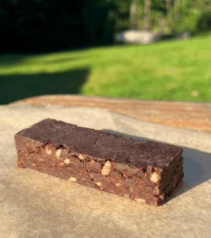 A chocolate brownie with nuts on a piece of parchment paper outdoors with grass and trees in the background.