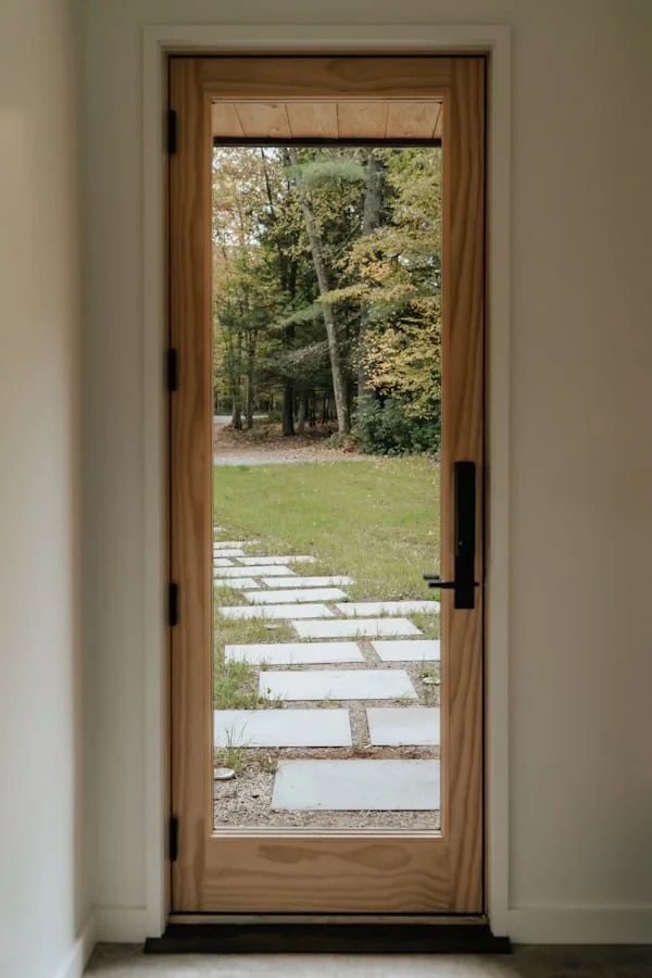 A wooden door with a glass pane, leading to a backyard with a pathway of concrete slabs and a wooded area with trees in the background.
