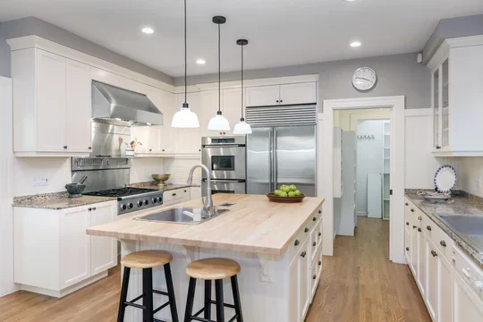 white cabinetry, pine hardwood floors, butcher board island with sink, pendant lighting, exhaust hood, and seating under waterfal