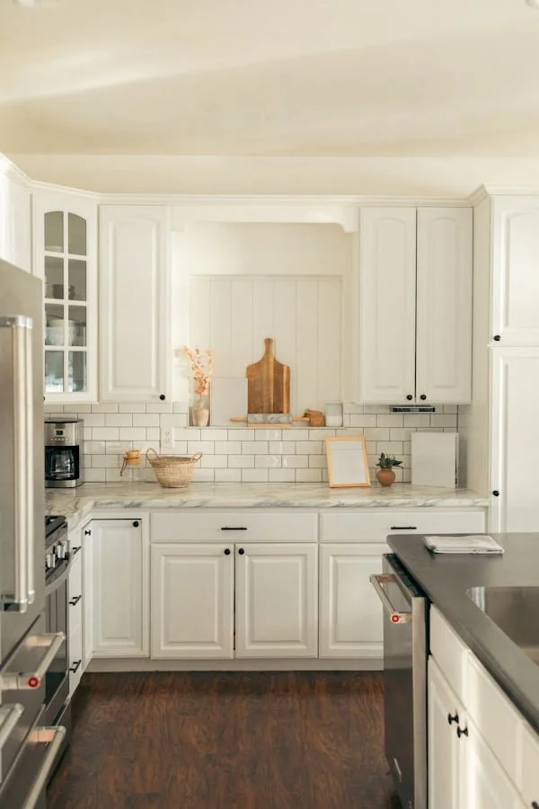 White kitchen with cabinets, a marble countertop, a white subway tile backsplash, and various decor items including a vase, a cutting board, and small plants.