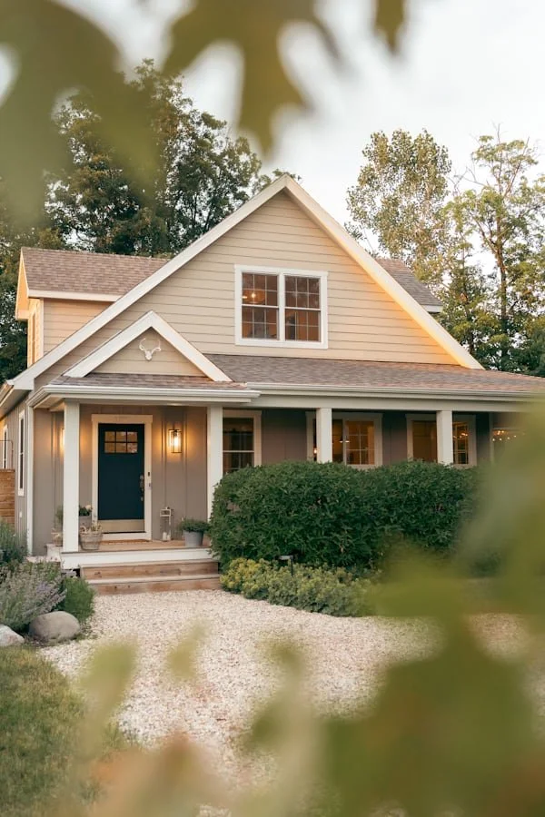 Front view of a beige two-story house with a gable roof, surrounded by greenery and trees, with a gravel pathway leading to the front door.