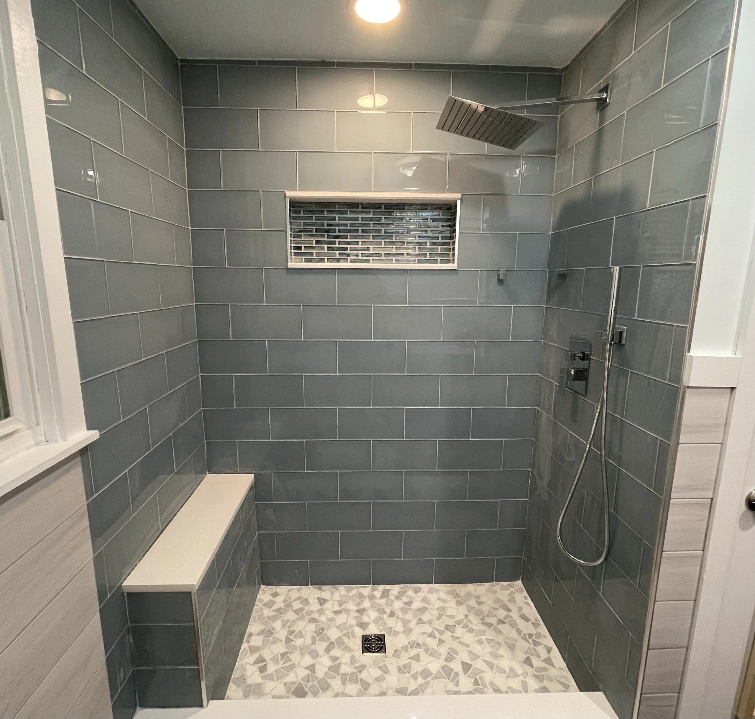 Custom tiled walk-in shower remodel in Horsham, PA featuring gray subway tile, rainfall showehead, built-in bench seat, glass mosaic niche, and pebble-style tie flooring
