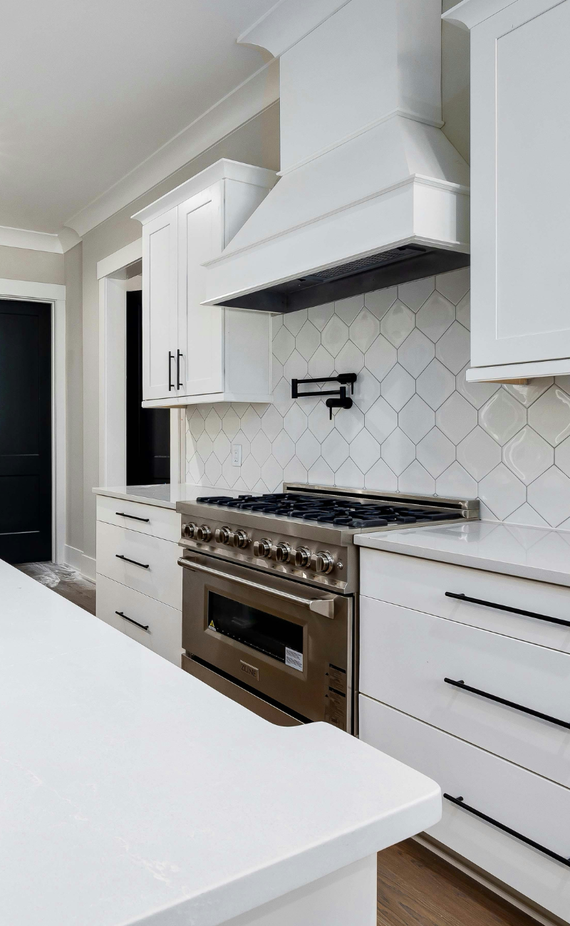 White kitchen with modern black handles, a stainless steel oven, a white countertop, and a black towel rack on the hexagon-tiled backsplash.