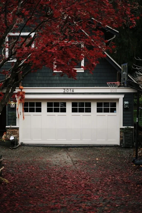 The image shows a house with a garage door, a basketball hoop on the side, and a large tree with vibrant red leaves in front of the house. There are Halloween decorations on the left side of the garage, and autumn leaves scattered on the driveway.