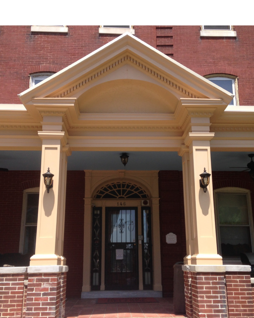 Front porch of a brick building with a cream-colored portico, columns, and decorative trim, featuring a black metal door with sidelights and a transom window, and two vintage-style lanterns flanking the entrance.