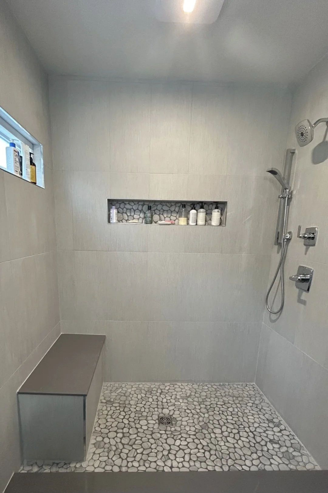Walk-in shower with large-format gray tile, pebble mosaic floor, built-in bench, and recessed shower niche