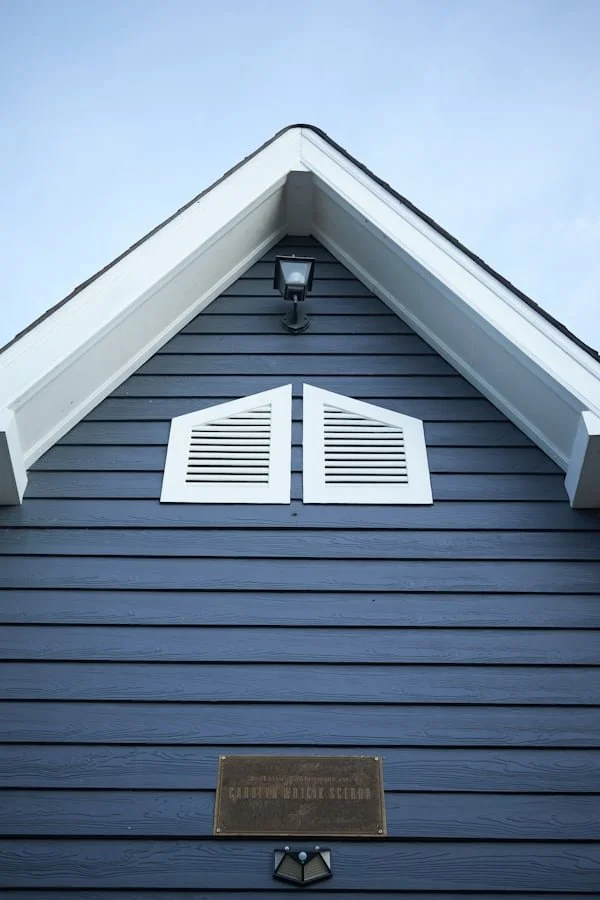 Close-up of the gable end of a blue house with white trim, window shutters, a light fixture, and a plaque.