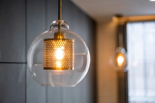 Close-up of a modern hanging glass pendant light with a decorative gold inner shade, glowing in a room with dark wall panels and a window.
