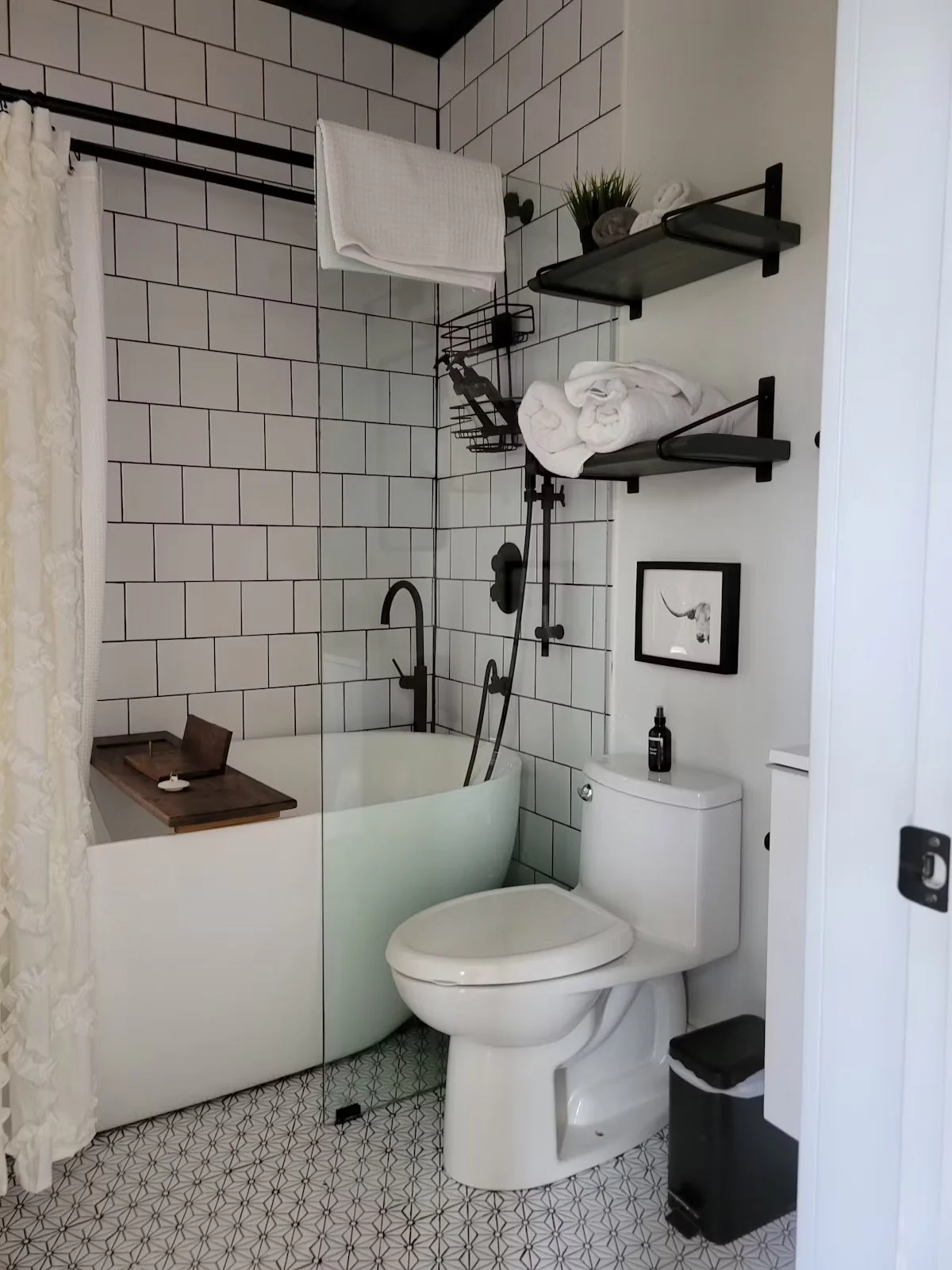 Bathroom with white tile walls, a bathtub with a wooden tray, a black-shower head and faucet, a toilet with a black soap bottle on top, black wall-mounted shelves with towels and a plant, a framed picture of a goat, and a small waste bin, with a black and white patterned tile floor.