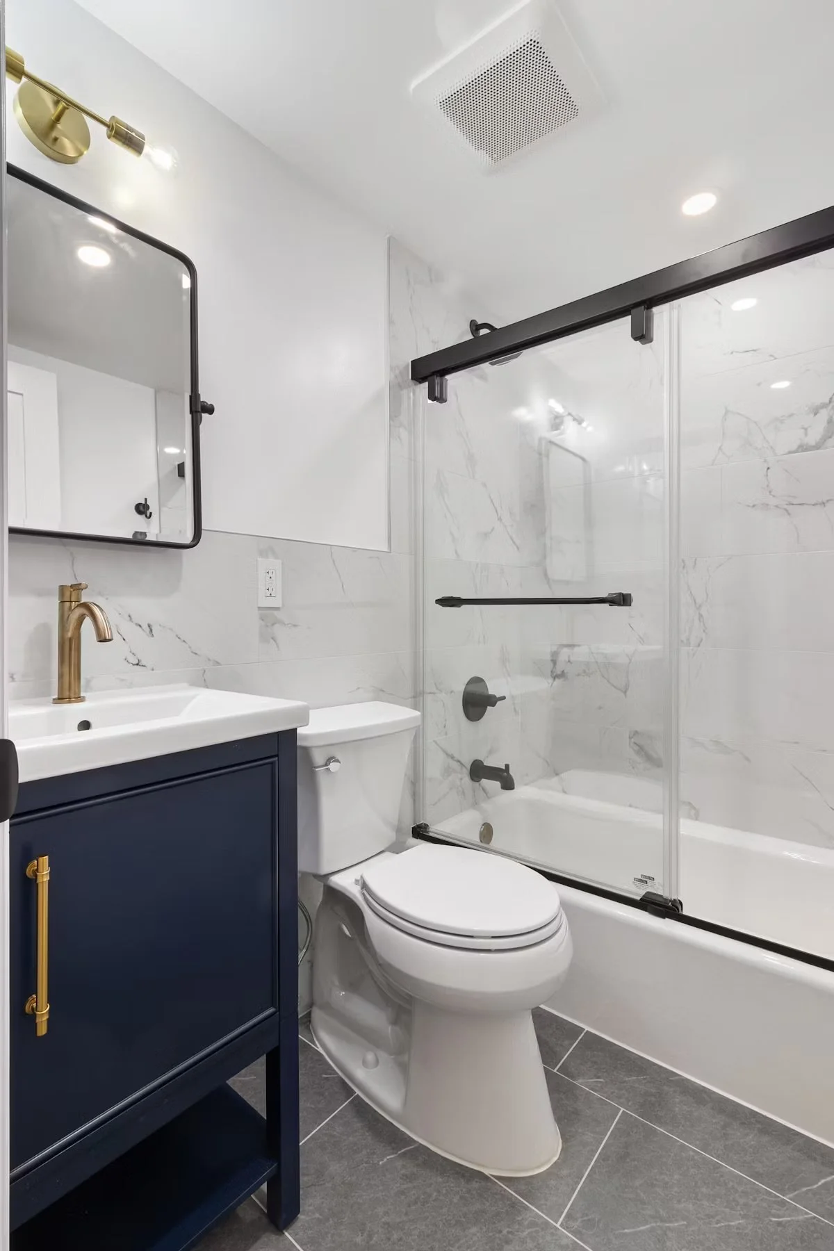 Modern bathroom with marble walls, a glass shower enclosure with black hardware, a white toilet, a navy blue vanity with gold accents, a gold faucet, a mirror, and a brass double light fixture.