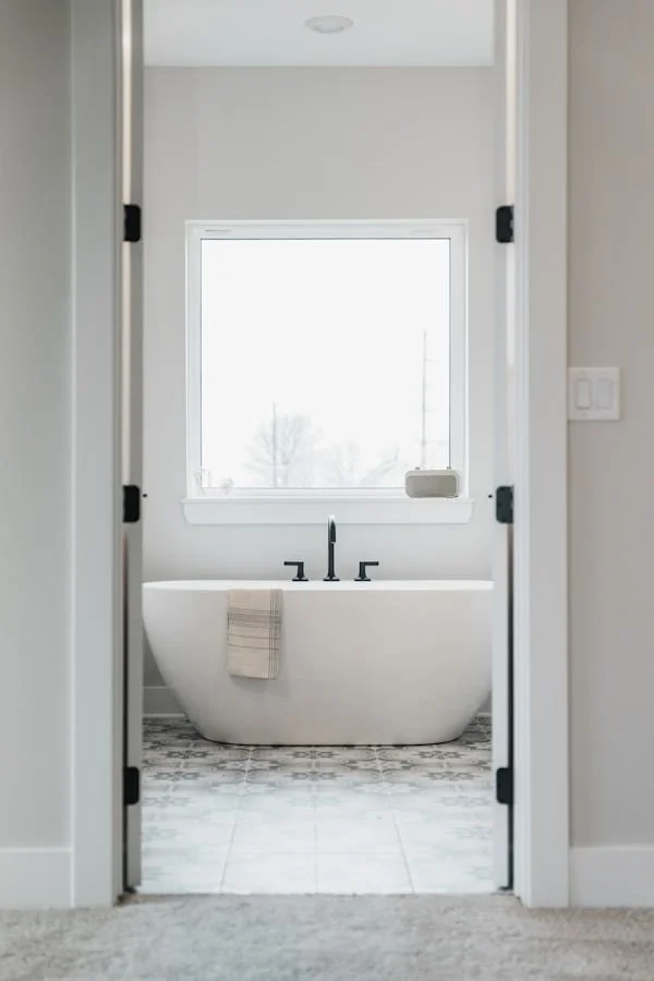 White freestanding bathtub with a beige towel hanging over the edge, positioned under a large window in a bright bathroom.