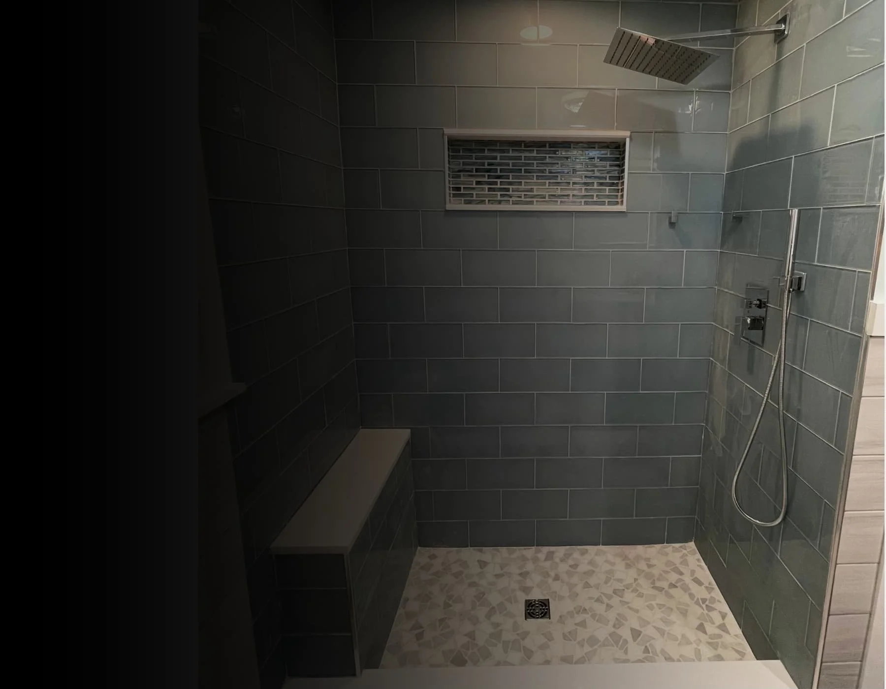 A modern shower with dark gray tiles on the walls, a stainless steel rain showerhead, a handheld shower wand, and a small rectangular window with a brick-patterned grille. The floor has small beige and gray mosaic tiles, and there is a drain in the center.