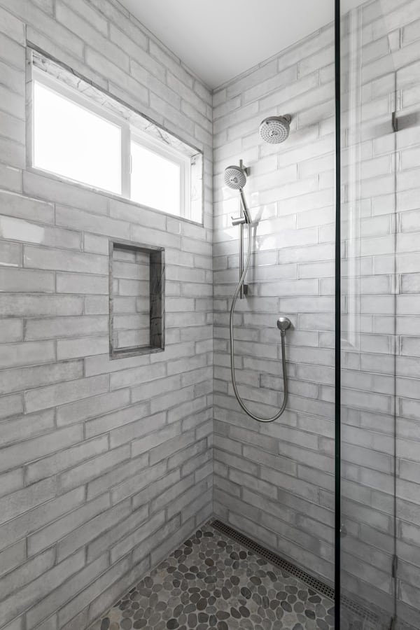 A modern walk-in shower with white brick tiles, a pebble stone floor, a rectangular niche in the wall, a small window for natural light, and a dual showerhead system.