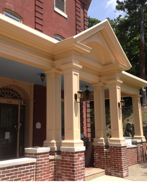 Porch with cream-colored columns, brick foundation, and decorative trim, attached to a red brick building, under a blue sky with trees in the background.