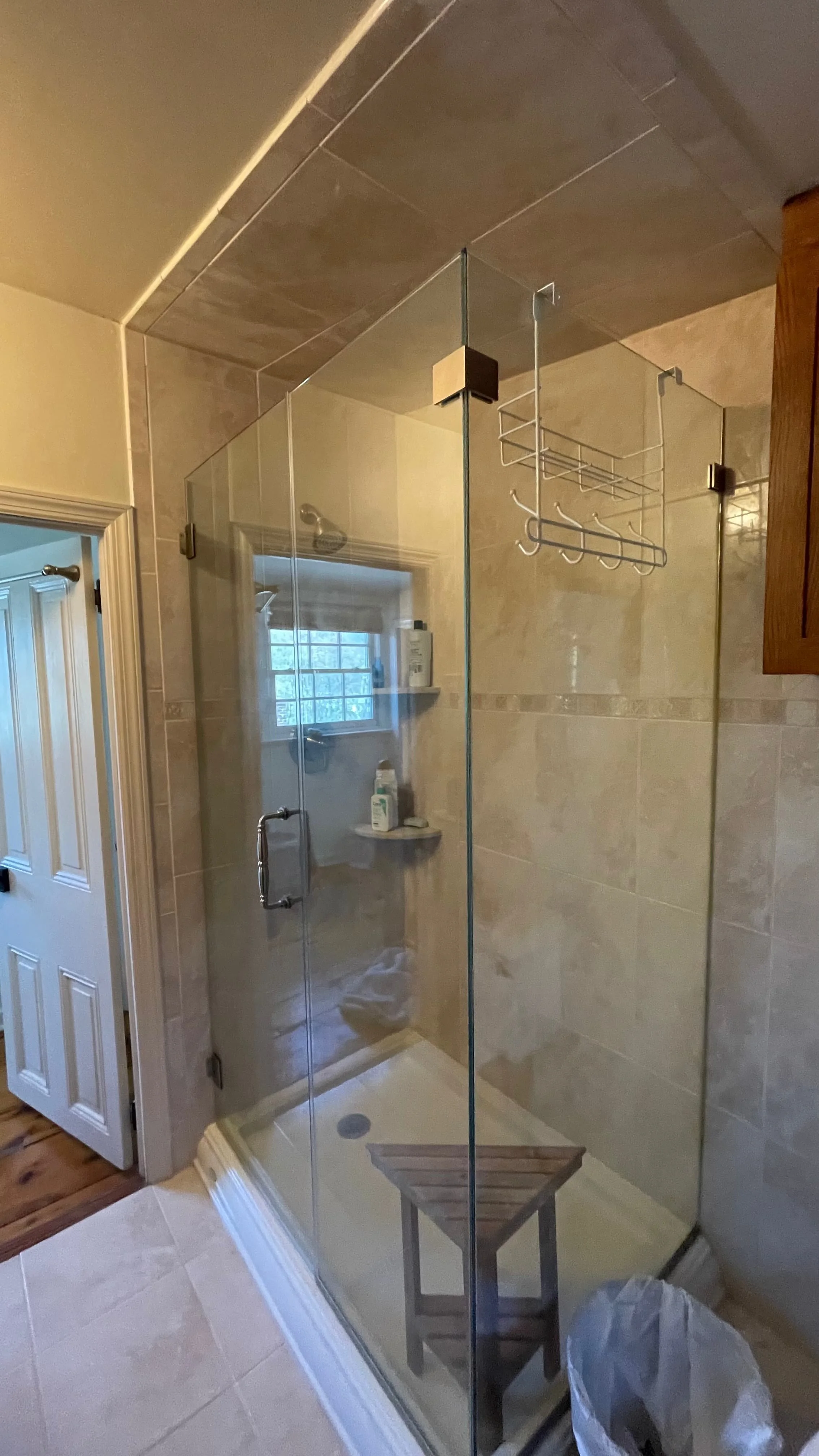 Existing frameless glass shower with tan tile walls, corner shelf, wall-mounted, shower head, and wood stool, before bathroom remodel in Hatboro, PA by JWD Contractors