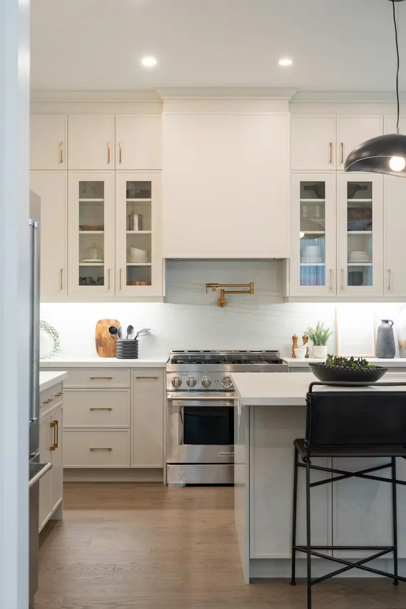 Modern white kitchen with stainless steel stove, glass-front cabinets, black bar stool, and minimal decor.