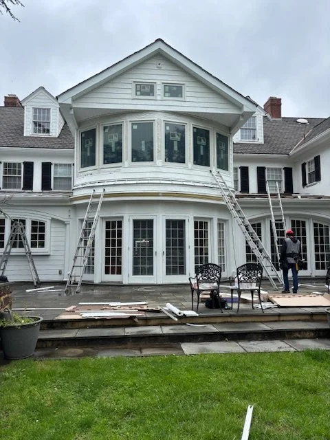 Construction workers installing or repairing windows on a large white house with a rounded bay window and a deck, ladders on both sides, and a person in a red helmet.