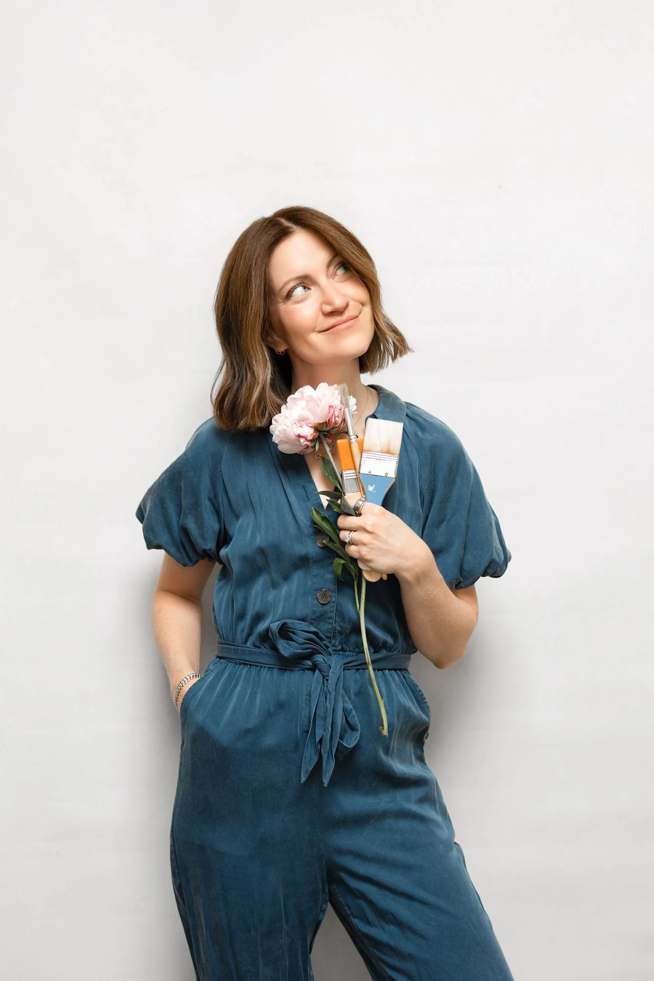 A woman with shoulder-length brown hair, wearing a denim jumpsuit, holds paint brushes and a pink peony flower against a plain white background, smiling and looking up.