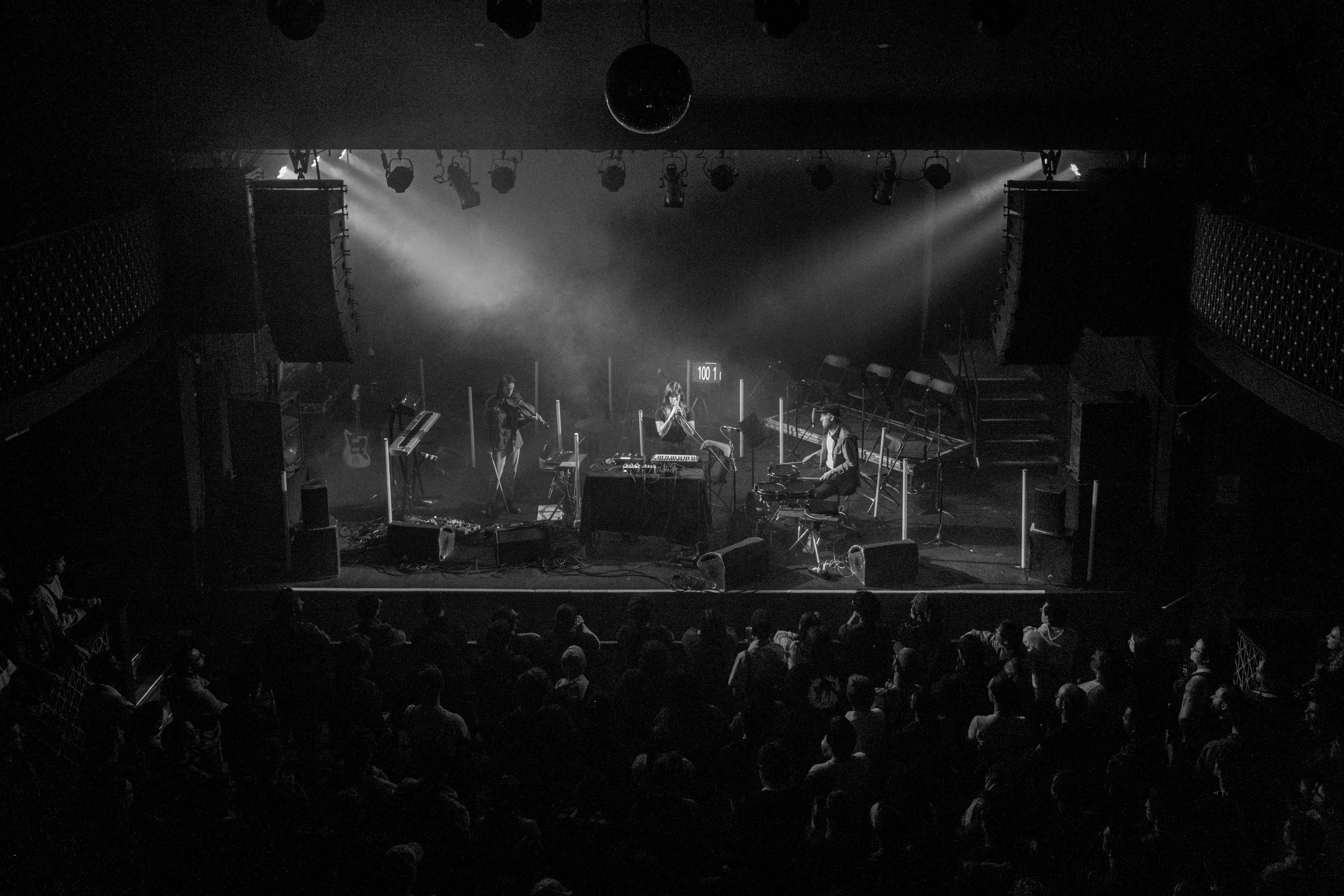 A musical performance on stage at a concert venue, featuring musicians with instruments including a keyboard, microphone, and drums, with an audience in front.