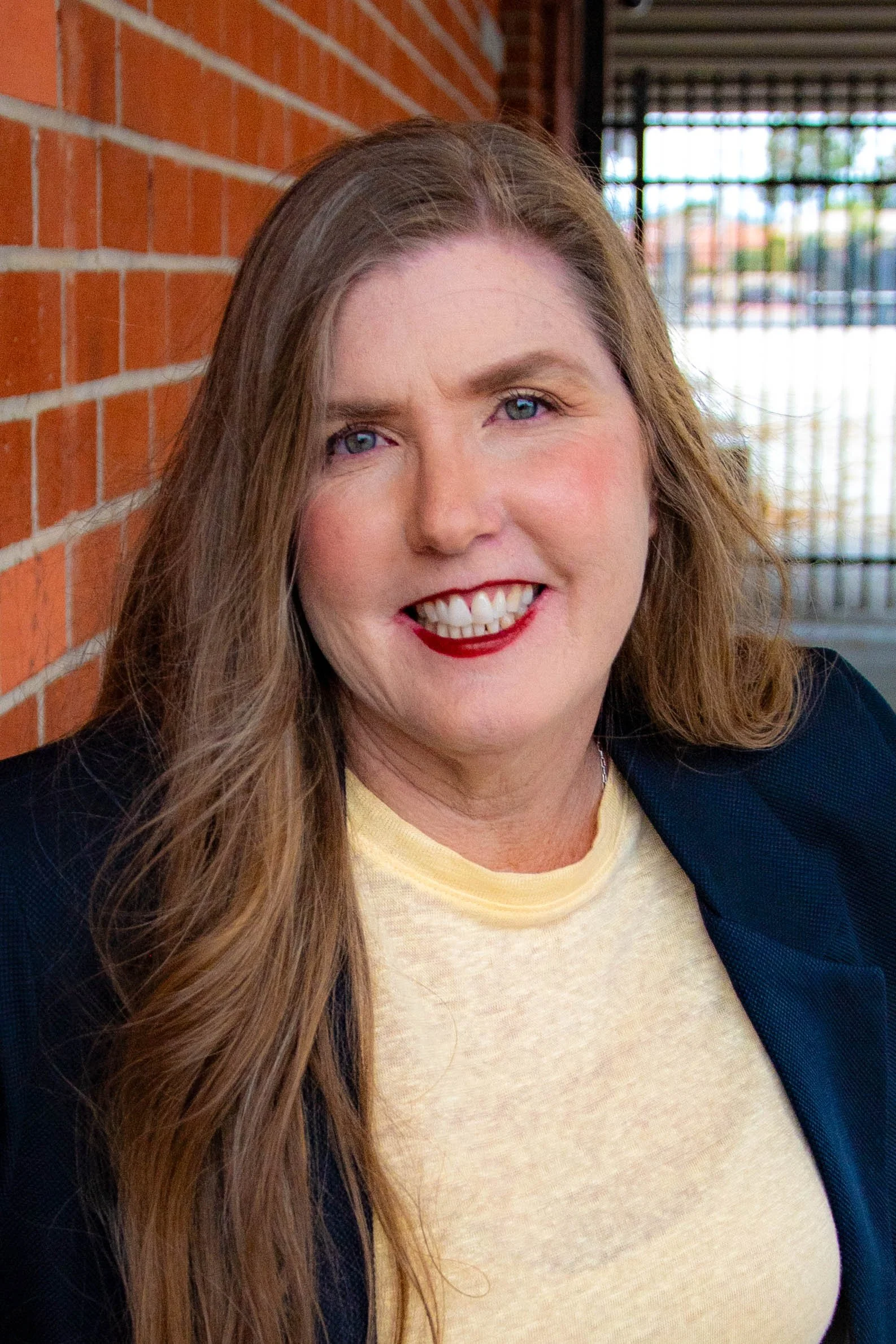 A woman with long brown hair, blue eyes, wearing red lipstick and a yellow t-shirt, smiling while leaning against a red brick wall with a window in the background.