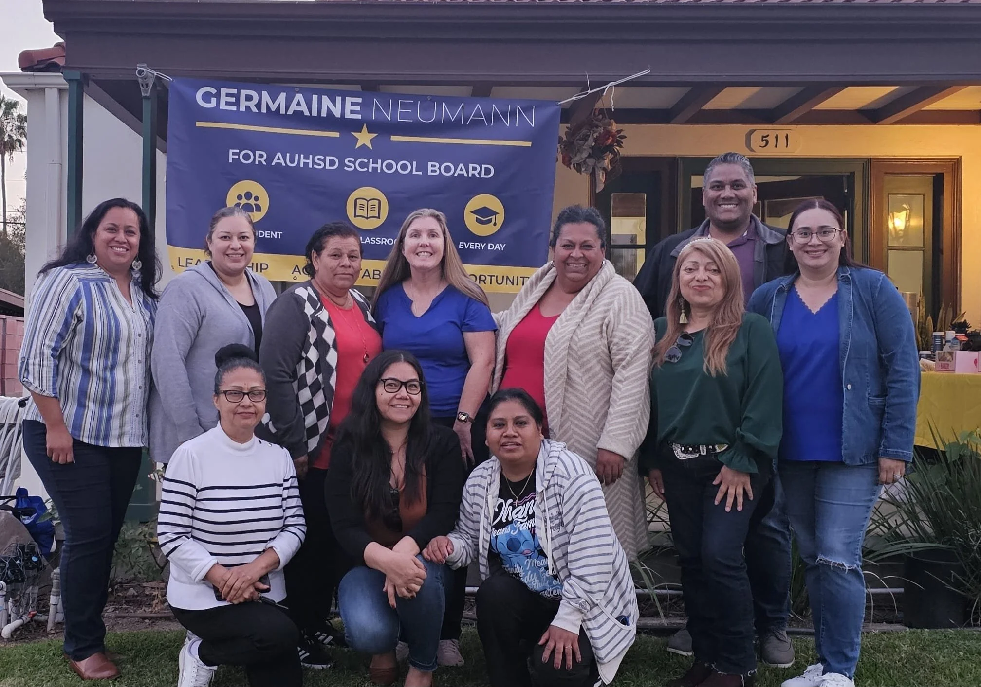 Group of diverse people standing in front of a house with a blue and yellow banner that reads 'Germaine Neumann for AUHSD School Board'.