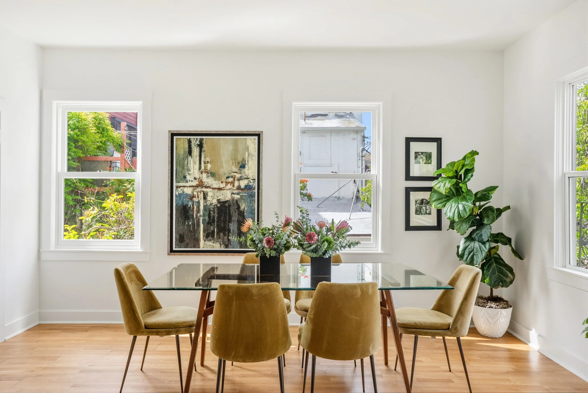 Dining room with six beige chairs around a glass top table, large plants and artwork, with three windows showing greenery outside.