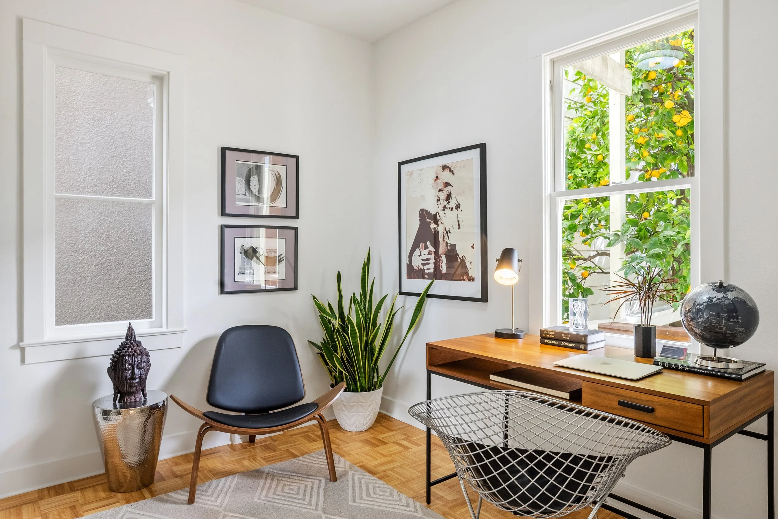 Bright home office with white walls, large windows showing lemon trees outside, a black office chair, a wooden desk with a globe, books, lamp, and plants, and framed artwork on the wall.