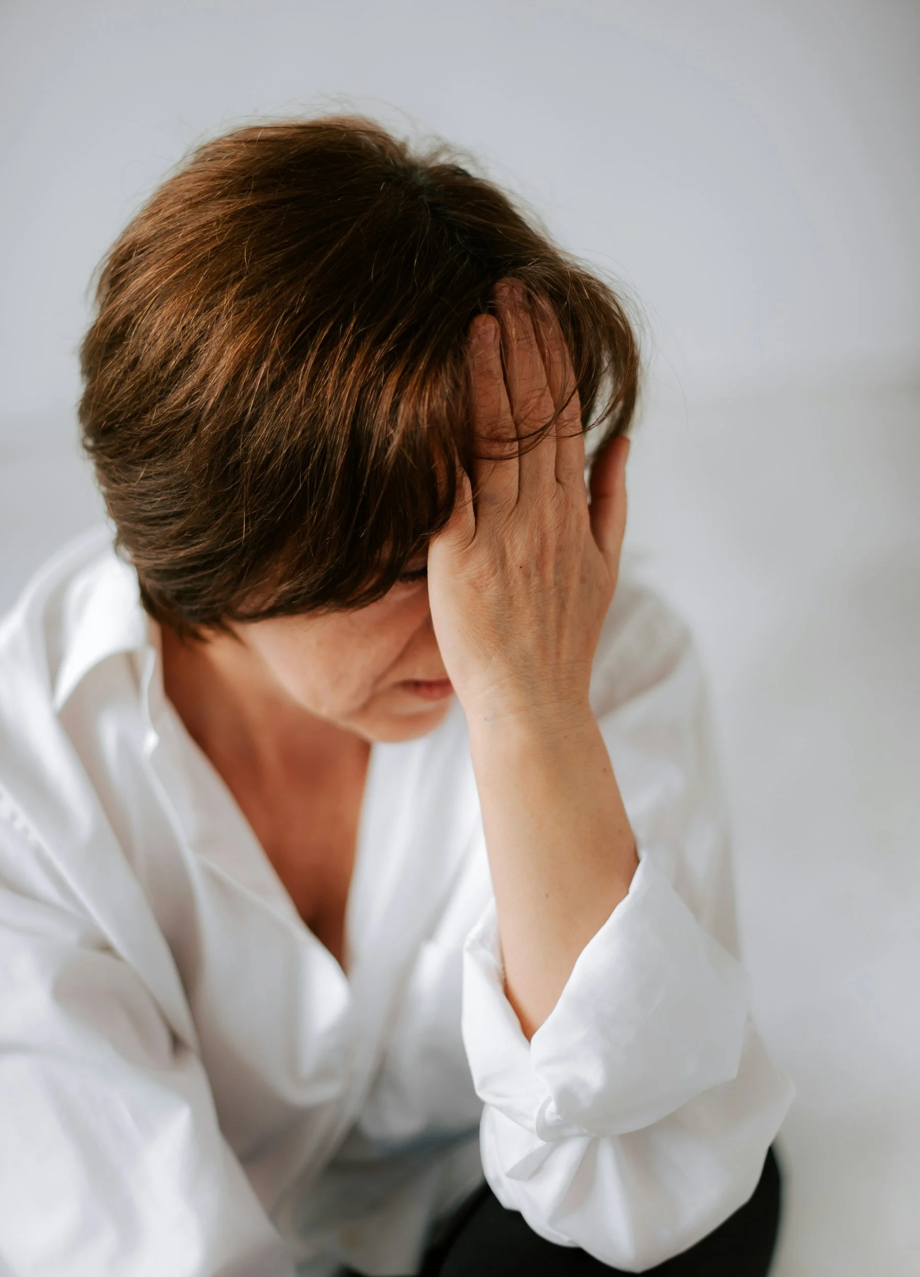 A woman with short brown hair, wearing a white blouse, sitting with her hand covering her face in a gesture of distress or frustration.