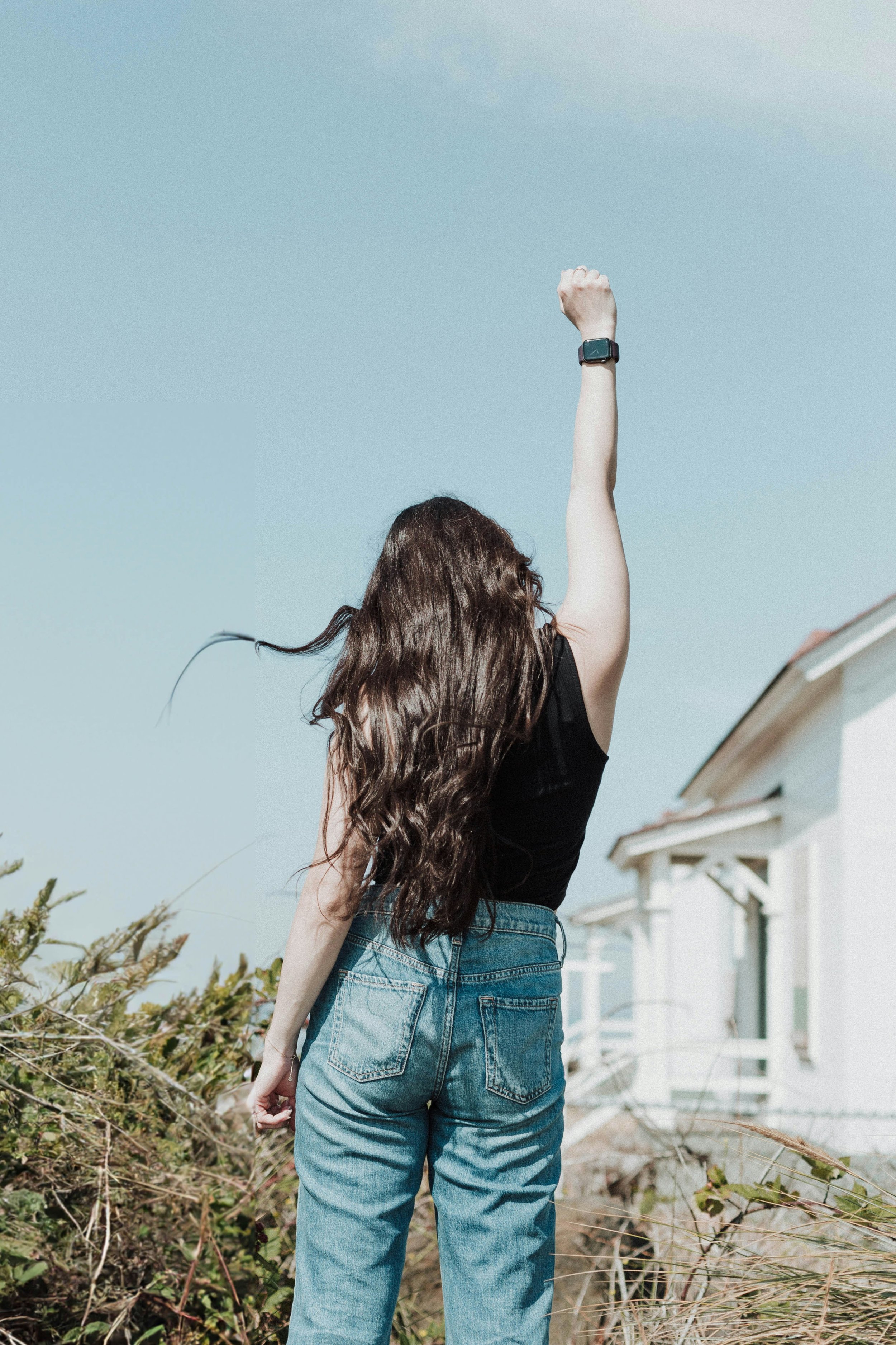 A woman with long wavy brown hair, wearing a black sleeveless top, high-waisted blue jeans, and a smartwatch on her left wrist, standing outdoors near some plants and bushes, raising her right arm in front of a row of white houses with porches, under a clear blue sky.