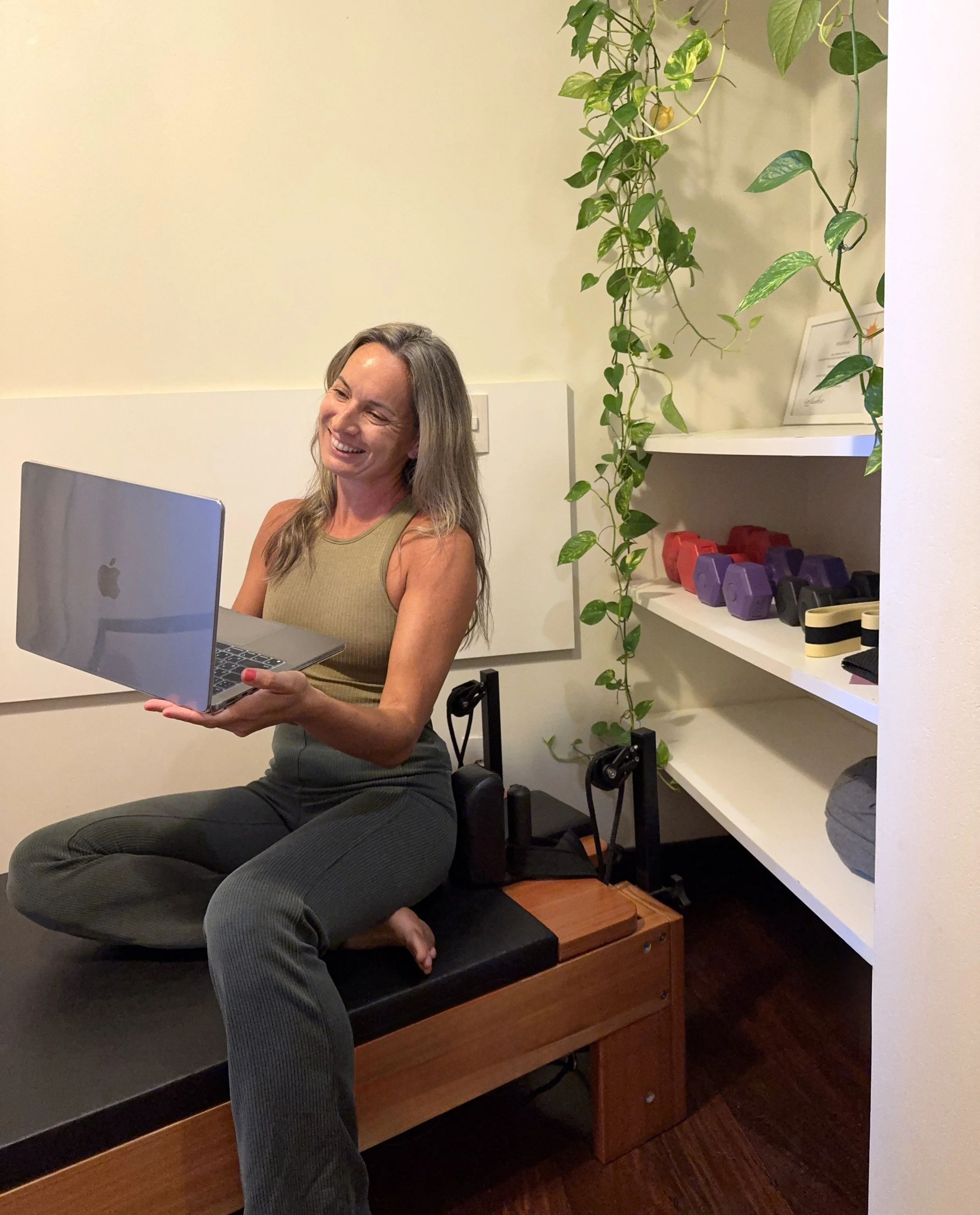 A woman sitting cross-legged on a wooden bench, smiling at a laptop, in a room with a white wall and shelves holding colorful dumbbells and decorative items, with green hanging plants.