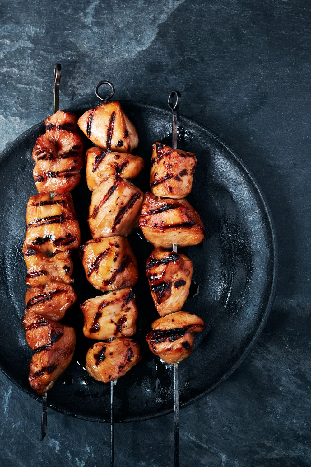 Skewers of grilled chicken pieces with grill marks on a black round plate.