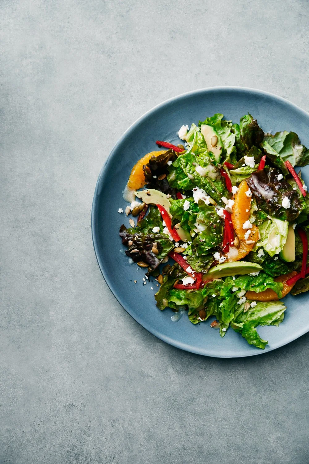 Fresh mixed green salad with orange slices, red peppers, nuts, and cheese on a blue plate